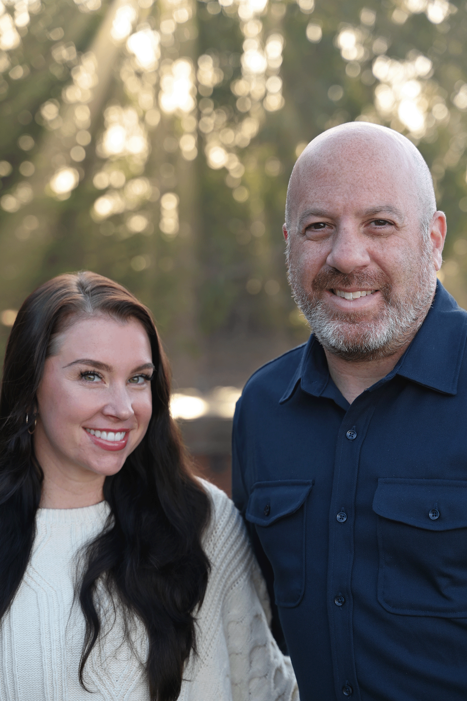 A woman and a man stand outdoors, smiling, with sunlight filtering through trees in the background.